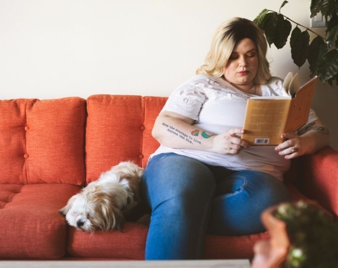 A pet sitter with blonde hair wearing a white shirt is sitting on an orange couch  reading a book with a small white dog beside her.